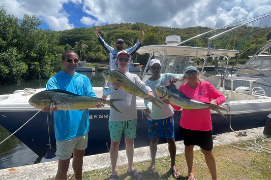 Four people in St.Croix holding large fish mahi by a boat on a dock with trees and sky in the background. St Croix Fishing Charter Mahi
